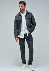 Hombre con chaqueta gris oscuro, camisa blanca, pantalones oscuros y zapatillas blancas, de pie con una mano en el bolsillo contra un fondo liso.