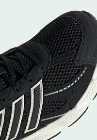 Chaussure de sport noire et blanche avec dessus en mesh, empiècements en suède, lacets noirs et trois bandes blanches sur le côté, vue en gros plan.