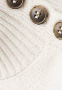 Details eines gestrickten cremefarbenen Pullovers mit gerippter Textur, drei runden, hellbraunen Knöpfen und sichtbaren Stichen auf dem Stoff.