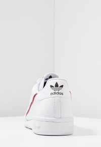 White Adidas sneaker viewed from the back, featuring red and navy stripes and the Adidas trefoil logo on the heel tab and sole.