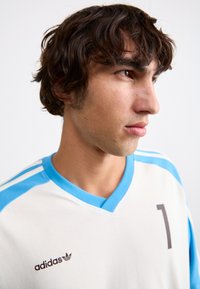 Maillot de sport blanc avec des accents bleu clair, doté d'un col en V, de trois bandes sur les épaules et d'un numéro « 1 » marron foncé. Logo Adidas sur la poitrine.