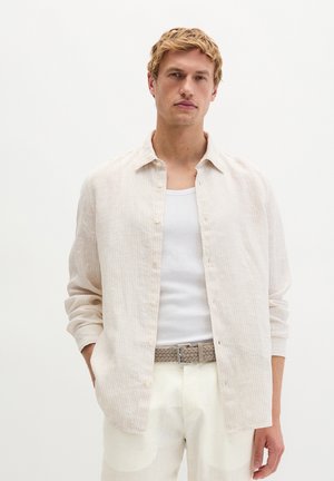 Junger Mann mit kurzem blondem Haar, der ein helles, gestreiftes Hemd mit Knöpfen, ein weißes Tanktop und weiße Hose mit geflochtenem Gürtel trägt, steht mit den Händen in den Taschen.