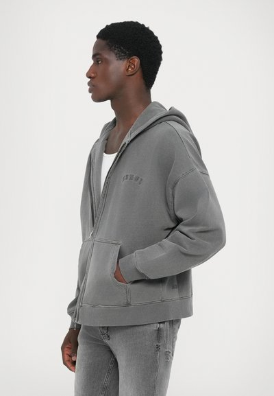 Grå zip-up hoodie i mjukt material, med en kenguruficka, justerbar huva och ribbade ärmar. Buren tillsammans med ljusgrå jeans.