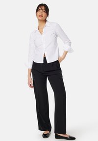 Camicia bianca con colletto, maniche lunghe arrotolate sopra i gomiti, abbinata a pantaloni neri a gamba larga su misura e ballerine nere.