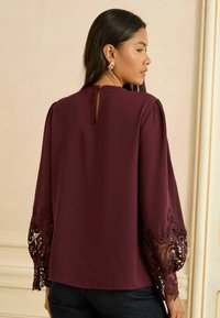 Vínová blúzka s dlhými rukávmi, zdobená zložitými čipkovými detailmi na manžetách a otvorom na chrbte. Hladká textúra látky.
