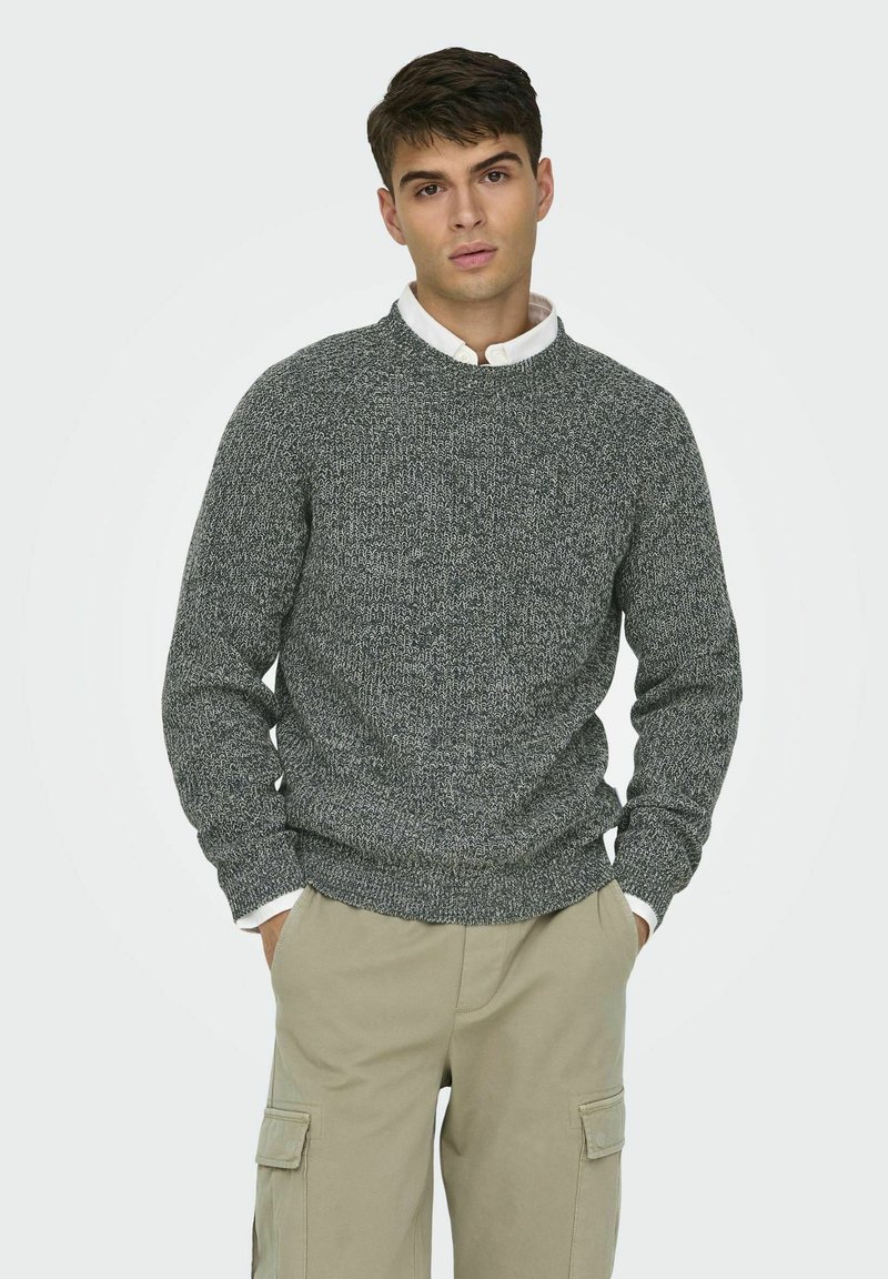 Grauer gestrickter Pullover mit Rundhalsausschnitt, strukturiertem Muster und gerippten Bündchen, getragen über einem weißen Hemd mit Kragen und kombiniert mit beigen Cargo-Hosen.