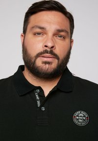 Czarny polo z kołnierzykiem, z okrągłą naszywką z logo i markowymi detalami na zapięciu. Krótkie rękawy z subtelnymi paskami.