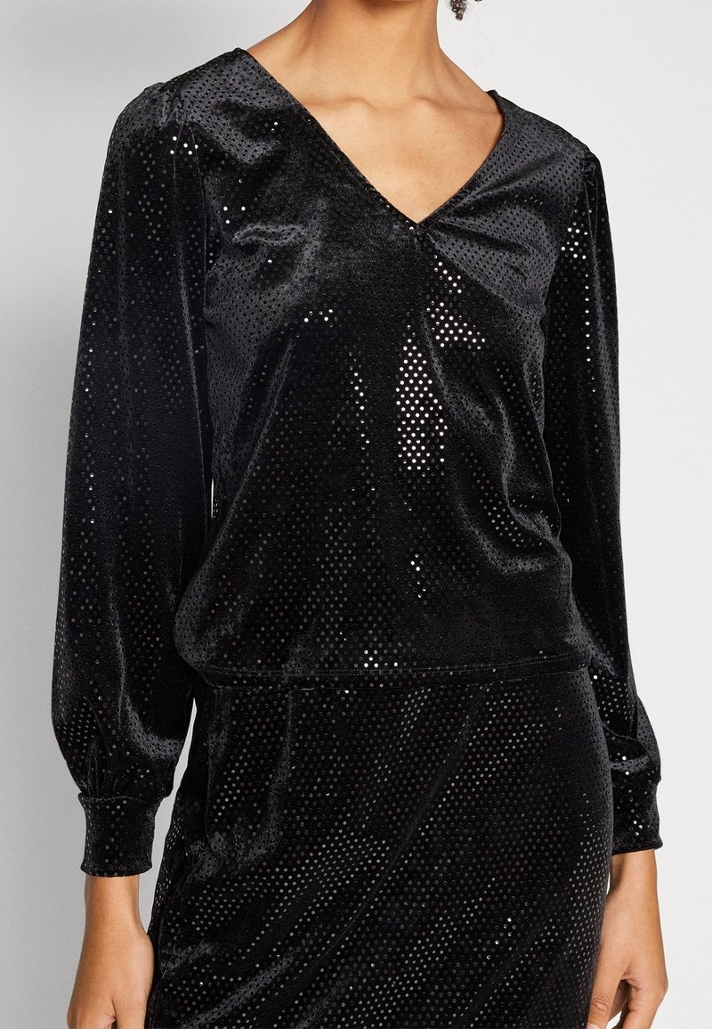 Svart sammetstopp med V-krage och långa puffärmar, med ett prickigt mönster för textur och en slät, reflekterande yta.