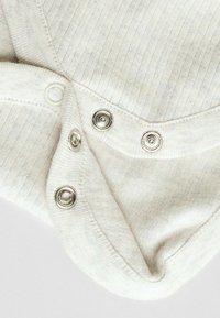 Long-sleeve light beige garment with a ribbed texture, featuring silver snap buttons and a rounded neckline.