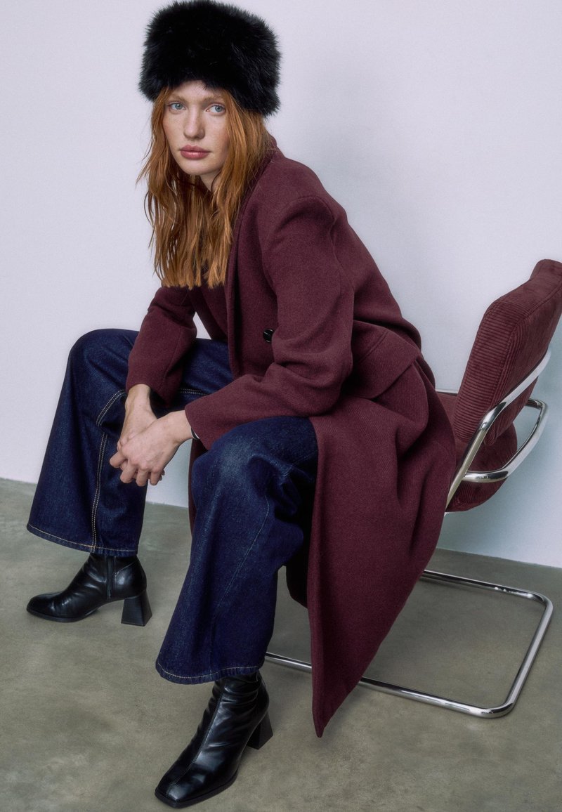 Cappotto di lana color mogano, jeans a gamba larga blu scuro e stivaletti neri con il tacco. Il soggetto indossa un cappello di pelliccia nero, seduto su una sedia argentata.