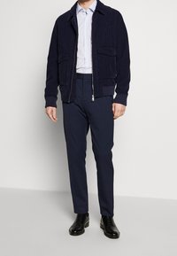 Marineblauwe corduroyjas met twee vo pockets, gecombineerd met een lichtblauw shirt en marineblauwe broek. Afgemaakt met zwarte veterschoenen.