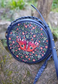 Ronde blauwe handtas met gevlochten riem, geborduurd met groene bladeren, roze bloemen en een grote rode vlinder in het midden.