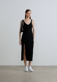 Čierne midi šaty bez rukávov s hlbokým výstrihom do V a bočným rozparkom, vyrobené z textúrovanej látky. Kombinované s bielymi teniskami a čiernou kabelkou na rameno.