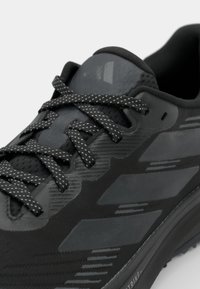 Scarpa sportiva nera con tomaia testurizzata, dettagli riflettenti e lacci con motivo. Design aerodinamico con suola ammortizzata per il massimo comfort.