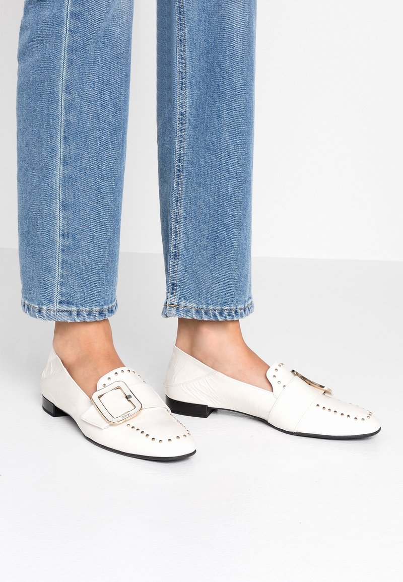Mocassins en cuir blanc avec des détails cloutés et une grande boucle rectangulaire. Petit talon et texture lisse, associés à un jean cropped bleu clair.