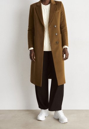 Abrigo largo marrón con cuello con muesca, botones cruzados, combinado con un suéter crema de punto trenzado y pantalones negros de pierna ancha, zapatillas deportivas blancas.