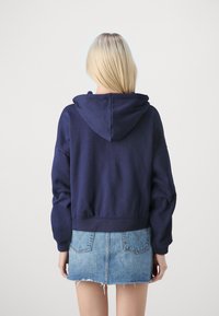 Donna bionda in piedi con la schiena rivolta verso la fotocamera, indossa un felpa blu navy e una gonna di jeans azzurri con orlo sfilacciato.