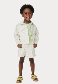 Chaqueta de mezclilla blanca y pantalones cortos con patrones florales coloridos, combinados con una blusa verde claro y sandalias amarillas con un acento de hoja.