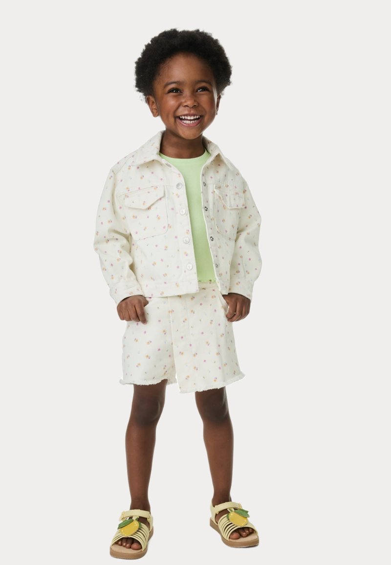 Chaqueta de mezclilla blanca y pantalones cortos con patrones florales coloridos, combinados con una blusa verde claro y sandalias amarillas con un acento de hoja.