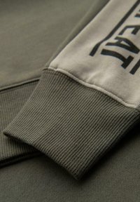 Tejido de sudadera verde con puños acanalados, que presenta un inserto beige con texto negro. Textura suave con un diseño casual.