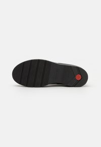 Semelle de chaussure en caoutchouc noir avec texture cannelée et logo circulaire rouge. Présente une surface lisse et des crampons bien définis pour l'adhérence.