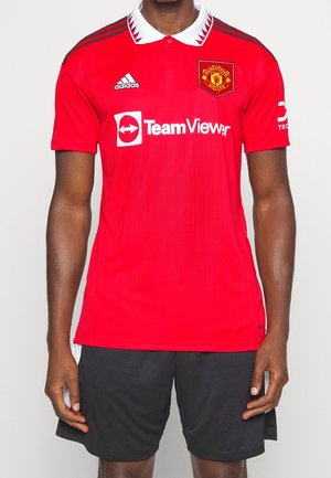 Mann trägt rotes Manchester United Fußballtrikot mit weißem Kragen und schwarzen Shorts, steht mit entspannten Armen an den Seiten.