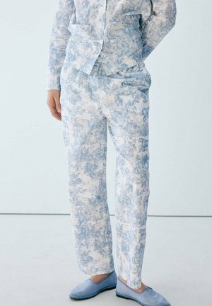 Persona con pantalones de pijama de color azul claro y blanco con patrón y zapatillas a juego, de pie con una mano en el bolsillo contra un fondo sencillo.