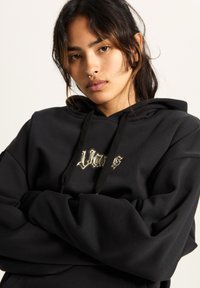 Schwarzer Hoodie mit einem großen, strukturierten Logo in heller Farbe. Ausgestattet mit einer Kordelzugkapuze und lockerem Schnitt, zeigt er glattes Material und gerippte Bündchen.