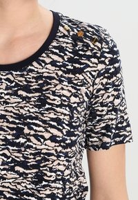 Camiseta de manga corta con un patrón abstracto en azul marino y crema, cuello redondo y botones de acento dorados en el hombro. Textura suave.
