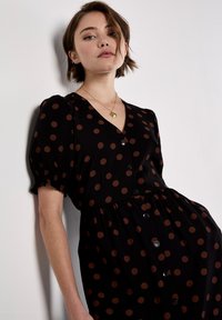 Zwarte jurk met korte pofmouwen, bruine stippen, V-hals, knoopsluiting aan de voorkant en een geplooide taille, met een zachte, gladde textuur.
