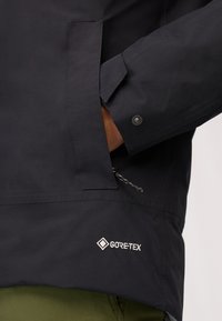 Giacca nera impermeabile con logo GORE-TEX, polsini regolabili, tasche con zip e finitura liscia opaca.