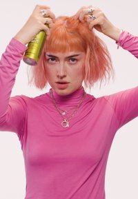 Une personne avec les cheveux courts rose portant un haut rose vaporise ses cheveux tout en les tenant, portant plusieurs bagues et des colliers superposés.