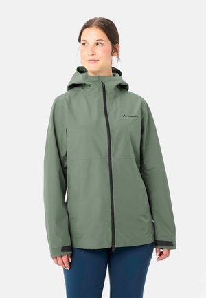 Vrouw met een groene regenjas met capuchon en een ritssluiting aan de voorkant, staand tegen een effen lichte achtergrond, terwijl ze opzij kijkt.