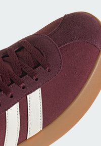 Burgundské semišové tenisky s bílými pruhy, kulatými tkaničkami a texturovanou gumovou podešví. Měkký svršek s hladkými akcenty.