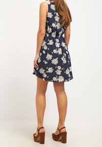 Námořnické květinové šaty s bílými sedmikráskami, bez rukávů, s úzkým pasem a rozšířenou sukní. Hnědé platformové podpatky, texturované dřevěné akcenty.