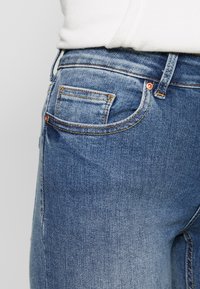 Primer plano de un bolsillo y una cinturilla de jeans de mezclilla azul con remaches de cobre, llevados con una blusa blanca metida por dentro.