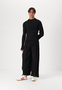 Svart plisserte langermet topp kombinert med vide plisserte bukser. Har en teksturert design. Modellen har på seg hvite joggesko med røde striper.