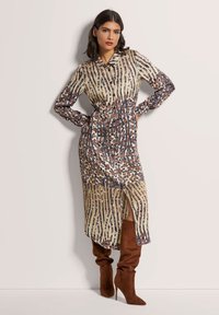 Langärmliges, kragenloses Kleid mit einem gemischten Tierdruckdesign, das vertikale Streifen in Gold und Braun aufweist, sowie einen seitlichen Schlitz. Braune kniehohe Stiefel.