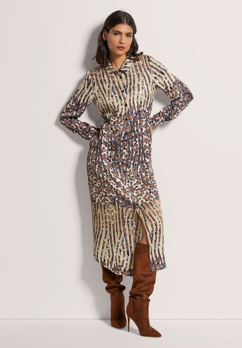 Langärmliges, kragenloses Kleid mit einem gemischten Tierdruckdesign, das vertikale Streifen in Gold und Braun aufweist, sowie einen seitlichen Schlitz. Braune kniehohe Stiefel.