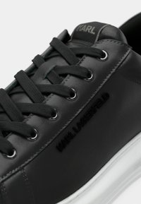 Sneaker din piele neagră, cu șnururi rotunde, un finisaj texturat și o talpă din cauciuc alb. Numele mărcii este embosat pe lateral.