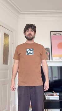 Mann mit dunklem Haar und Bart, trägt ein braunes T-Shirt und dunkle Hose, steht in einem Raum mit Fernseher, Büchern und gerahmten Kunstwerken an weißen Wänden.