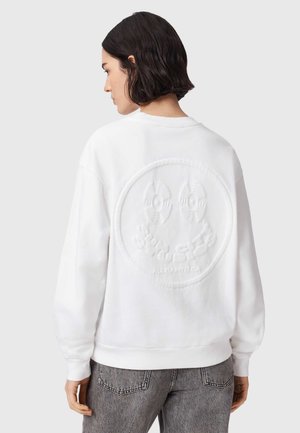 Kobieta o krótkich ciemnych włosach, ubrana w białą bluzę z tłoczonym okrągłym wzorem na plecach oraz szare dżinsy, zwrócona w lewo.