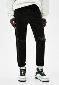 Černé džíny z denimu s roztrhanými koleny a vyhrnutými nohavicemi, doplněné bílými, zelenými a černými kotníkovými teniskami.