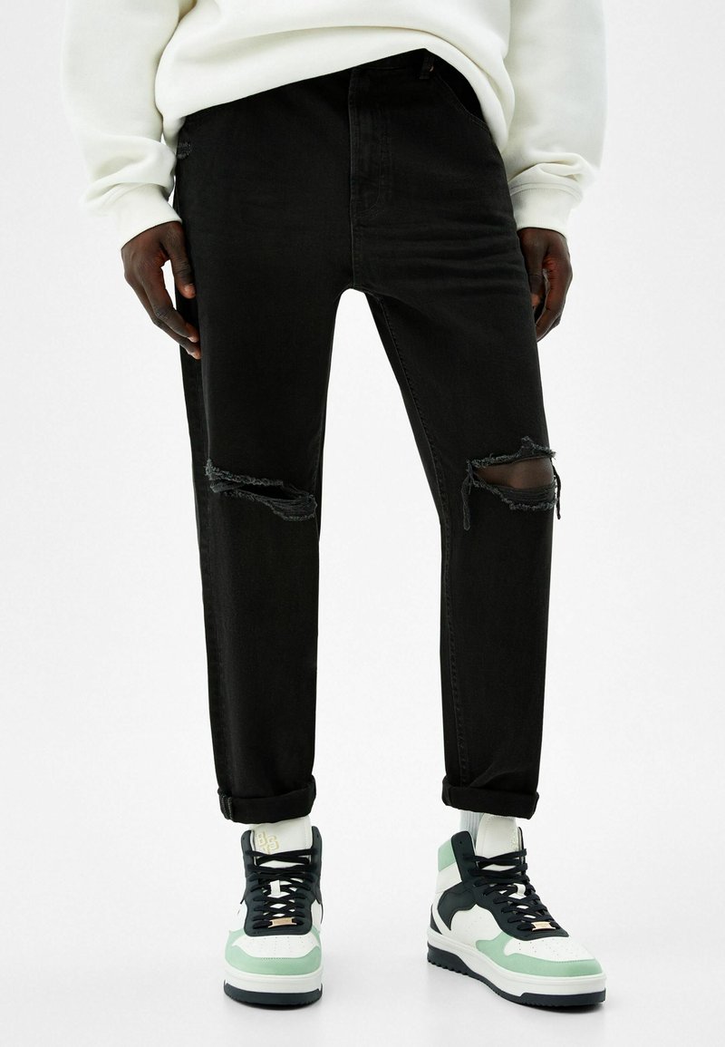 Černé džíny z denimu s roztrhanými koleny a vyhrnutými nohavicemi, doplněné bílými, zelenými a černými kotníkovými teniskami.