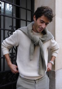 Young man with curly hair wearing a beige knit sweater and gray sweater draped over shoulders, standing near a black wrought iron gate.
