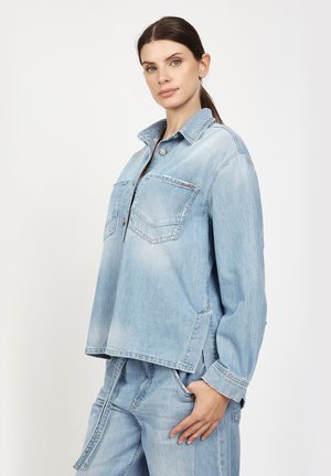 Woman with brown hair in a ponytail wearing a light blue denim jacket and matching jeans, standing with one hand in pocket against white background.