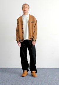Brauner Cardigan mit schwarzen Streifen, weißem T-Shirt, schwarzen Hosen und sandfarbenen Sneakern. Der Cardigan hat eine Knopfverschluss und ein Logo auf der Vorderseite.