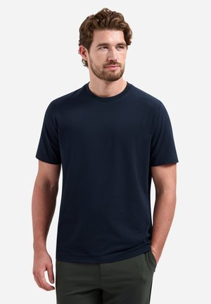 Man with light skin and curly brown hair wearing a plain dark navy blue short-sleeve T-shirt against a white background.