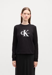 Juoda ilgo rankovės džemperis su baltu Calvin Klein logotipu. Pagamintas iš minkštos medžiagos, turi rausvas rankogalius ir laisvą siluetą.