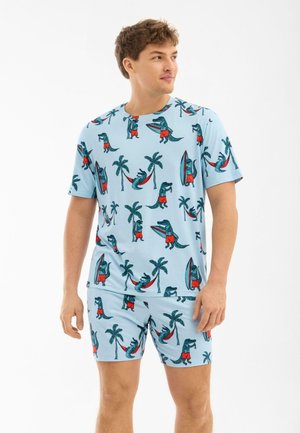 Mies pukeutunut vaaleansiniseen yhteensopivaan paitaan ja shortseihin, joissa on leikkisä alligaattori-, palmupuu- ja surffilautaprintti.